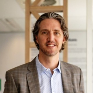 Man in a gray blazer smiles in a modern office environment with a light background.