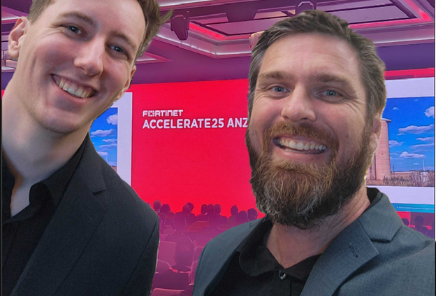 Two smiling men in suits pose for a selfie at a conference with a colorful background.