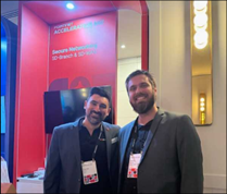 Two men in blazers smiling at a booth with a red backdrop displaying "Accelerating."