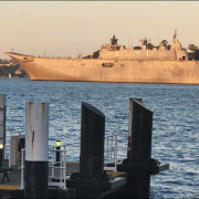 Large naval ship sails in calm waters, with docks and pilings in the foreground.